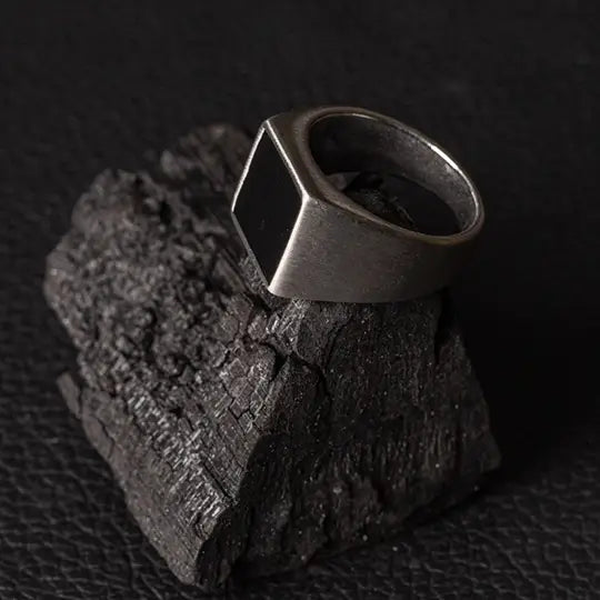 Anel de aço Vittorio com design geométrico, repousando sobre um carvão texturizado em fundo preto.