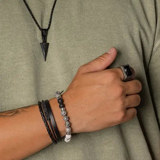 Pulso masculino com mix de pulseiras de couro e pedras e anel de aço geométrico, complementado por colar com pingente de flecha.