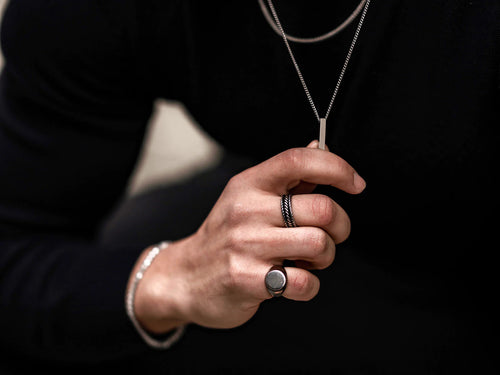 Mão de homem com dois anéis, um trançado no dedo médio e outro com pedra escura no dedo anelar, segurando delicadamente um colar com um pingente retangular prateado, contra um fundo escuro e desfocado.