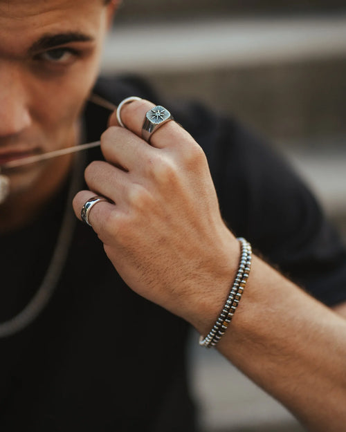 Um homem com expressão focada segura delicadamente um colar de corrente prateada, exibindo anéis decorativos no dedo indicador e anelar e uma pulseira de contas no pulso, tudo contra um fundo suavemente desfocado.