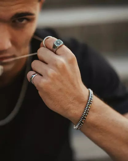Homem desfocado ao fundo ajustando colar enquanto exibe anéis prateados com detalhes no dedo indicador e anelar e uma pulseira de contas no pulso.