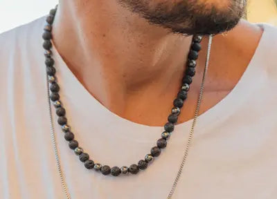 Close-up de um homem usando uma camiseta branca com um colar de pedras vulcânicas e uma corrente prateada fina ao redor do pescoço.