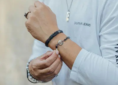 Pessoa usando uma camiseta branca com a logo 'Calvin Klein Jeans', complementada por pulseiras de couro trançado e uma com pedras naturais brancas, ajustando-as no pulso.