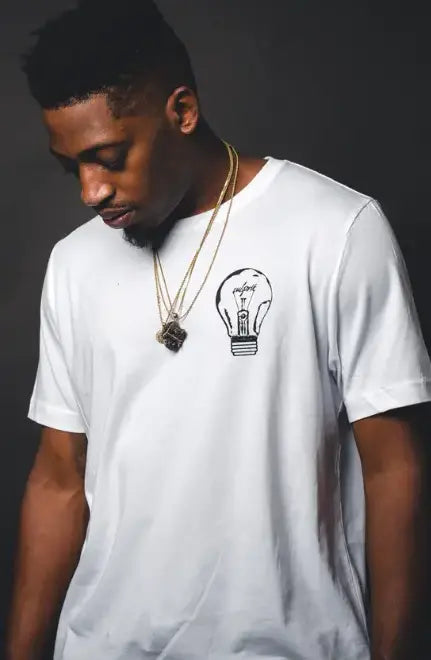Homem olhando para baixo vestindo uma camiseta branca com um desenho de lâmpada no peito, usando vários colares com um pendente de leão.