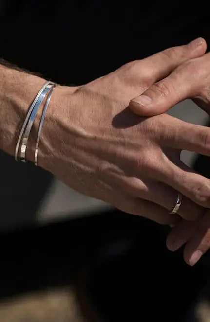 Mão masculina com detalhes visíveis da pele e veias, exibindo uma aliança simples no dedo anelar e uma pulseira de prata, contra um fundo desfocado.