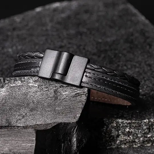 Pulseira de couro genuíno HeFesto preta com fecho magnético fosco e detalhes trançados, em close-up sobre uma superfície de carvão e pedra de mármore texturizada no fundo.