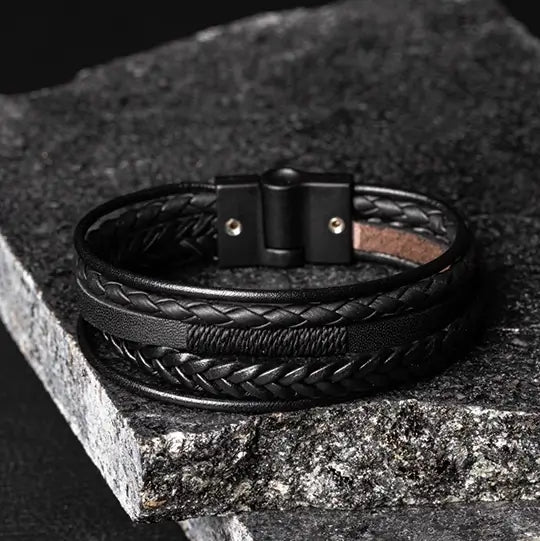 Pulseira de couro preto trançado com fecho magnético, elegantemente disposta sobre uma pedra rústica.
