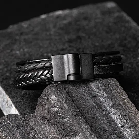 Pulseira de couro preto com trançado e fecho magnético metálico, repousando sobre uma superfície de madeira envelhecida e pedra.