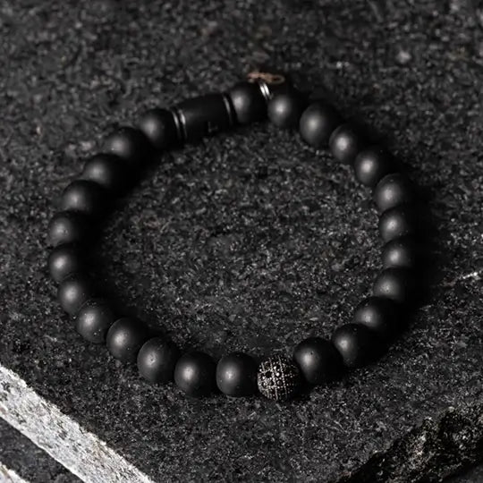 Elegante pulseira de contas pretas com uma peça central incrustada de cristais, colocada sobre uma superfície texturizada de pedra escura.
