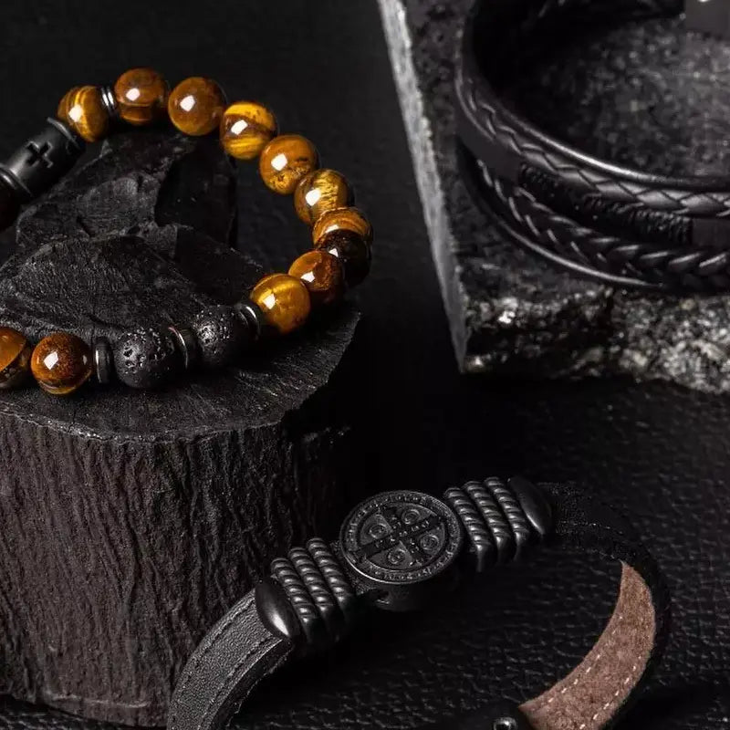Variedade de pulseiras de design sofisticado em close-up sobre uma superfície preta, destacando uma pulseira de contas âmbar e lava, uma pulseira trançada de couro preto e uma pulseira de couro com detalhe de fecho metálico com símbolos.